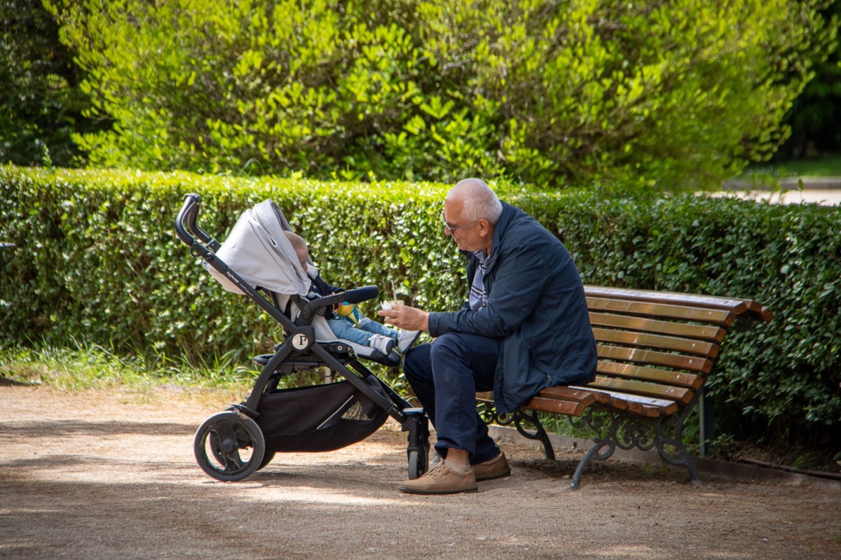 A grandfather on a park bench, leaning in to his grandchild sitting in a pushchair.